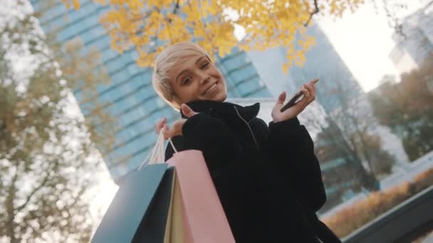 Junge Frau steht mit Einkaufstüten und Smartphone im Park und lächelt — Stockvideo