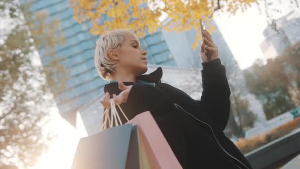 Młoda kobieta zakupoholiczka stojąca w parku z torbami na zakupy i korzystająca ze smartfona do robienia selfie — Wideo stockowe