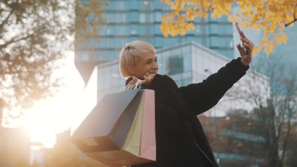 Junge Frau steht mit Einkaufstüten im Park und macht Selfie mit Smartphone — Stockvideo