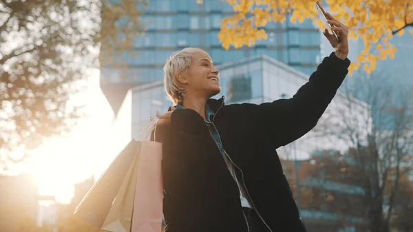 Junge Frau steht mit Einkaufstüten im Park und macht Selfie mit Smartphone — Stockfoto