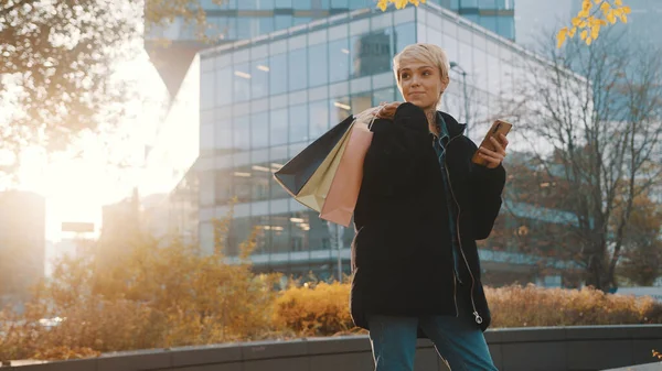 Winkelen in de herfst. Jonge blonde vrouw met behulp van telefoon in het park, terwijl het houden van boodschappentassen over de schouder — Stockfoto