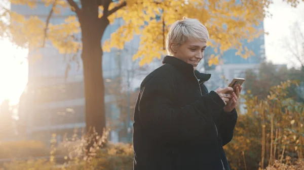 Retrato de jovem caucasiana usando telefone no parque da cidade em frente ao prédio de escritórios no outono — Fotografia de Stock