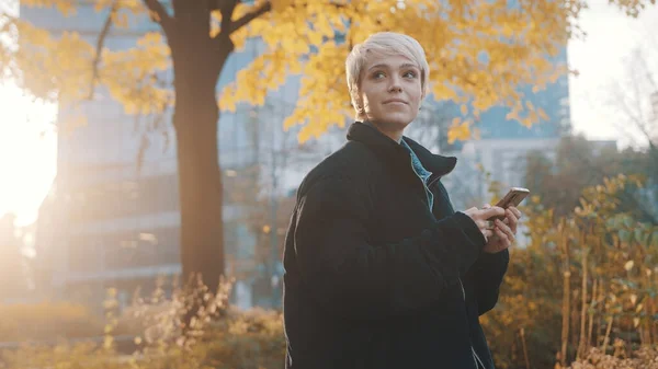 Retrato de una joven mujer caucásica usando teléfono en el parque de la ciudad frente al edificio de oficinas en otoño — Foto de Stock