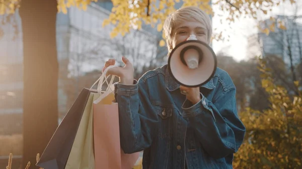Junge blonde Frau mit Einkaufstüten kündigt im Herbst mit Lautsprecher Verkaufssaison im Stadtpark an — Stockfoto