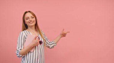 Young woman with striped shirt and notebook in hands pointing on the empty space, pink background