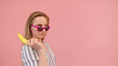 Young funny woman with pink sunglasses holding banana as pistol. Copy space