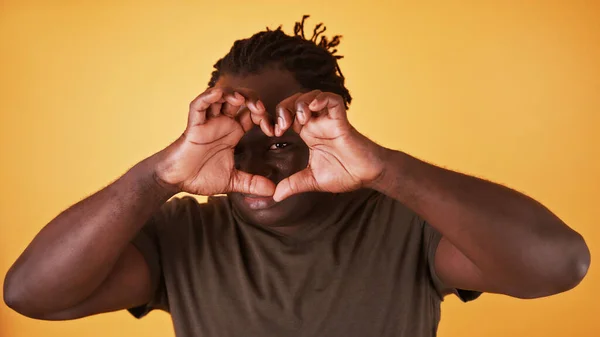 Homem negro afro-americano fazendo forma de coração com as mãos e olhando através dele. Amor romance e conceito de relacionamento — Fotografia de Stock