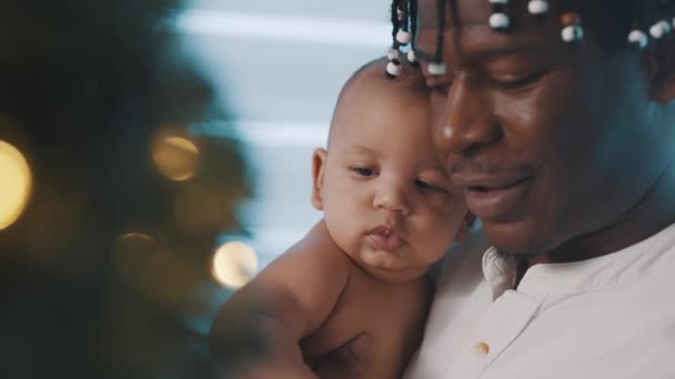 Gros plan, bébé nouveau-né et père câlins devant le sapin de Noël avec des lumières dorées — Video