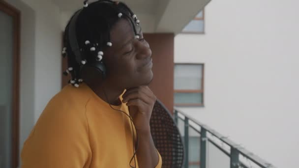 Porträt eines afrikanisch-amerikanischen schwarzen Mannes mit Dreadlocks, der per Headset auf dem Balkon Musik hört — Stockvideo