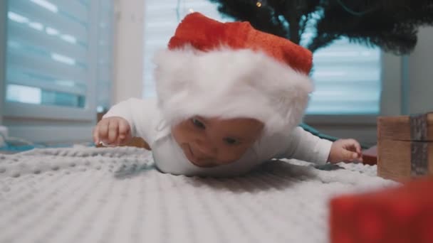 Schattige donkere huid baby met santa hoed omgeven door geschenken genieten van buiktijd onder de kerstboom — Stockvideo