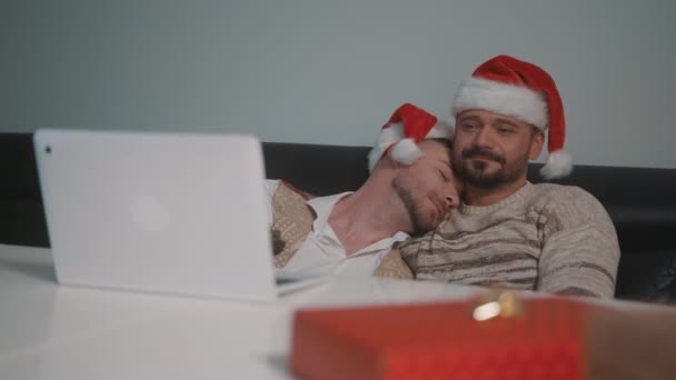 Szczęśliwy gej mężczyzna para z Santa Claus kapelusze przytulanie i oglądanie film na w Wigilia — Wideo stockowe