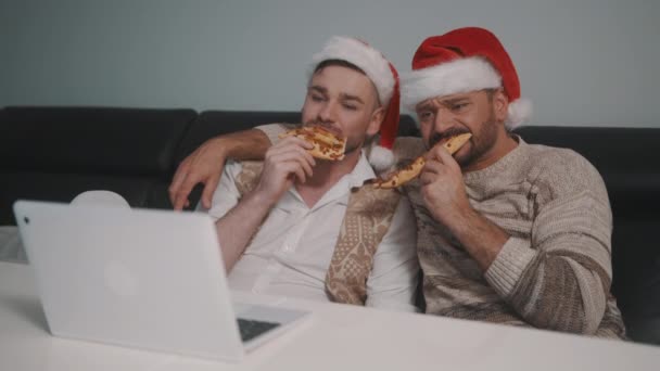 Ευτυχισμένο ζευγάρι ομοφυλόφιλων με καπέλα Αϊ Βασίλη που βλέπουν ταινίες στο λάπτοπ και τρώνε πίτσα την παραμονή των Χριστουγέννων — Αρχείο Βίντεο