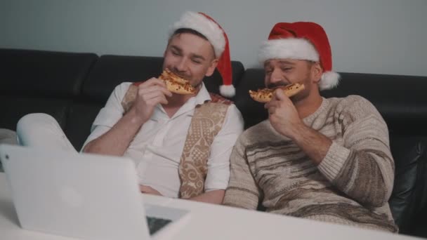 Noel Baba şapkalı mutlu eşcinsel çift dizüstü bilgisayarda film izliyor ve pizza yiyorlar. Aynı cinsiyette Noel kutlaması — Stok video