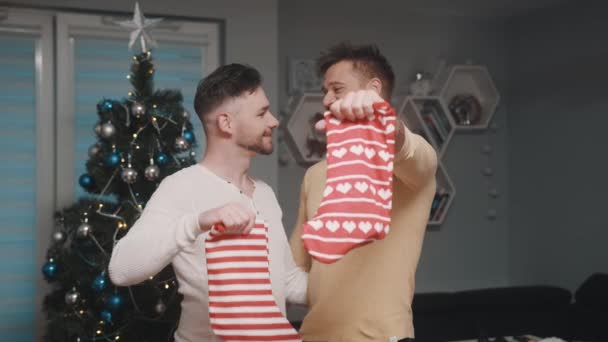 Радостная гей-пара в любви держит рождественские чулки перед елкой. Сочельник и гомосексуальные отношения — стоковое видео