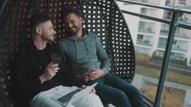 年轻漂亮的一对同性恋情侣在阳台上悠闲自在地荡漾，一边喝啤酒，一边在笔记本电脑上浏览社交媒体 — 图库视频影像