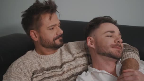 Закрывай. Два гея в повседневной одежде сидят на диване, обнимаются и смотрят телевизор дома — стоковое видео