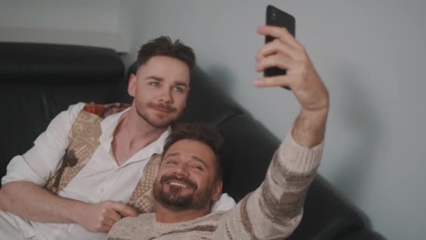 Ανδρικό ρομαντικό γκέι ζευγάρι χαλαρώνει στον καναπέ. Ο φίλος του φιλάει τον συνεργάτη του στο κεφάλι. Λήψη selfie με smartphone — Αρχείο Βίντεο