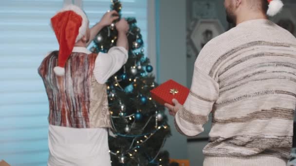 Achteraanzicht van de jonge Kaukasische man met Santa hoed versieren van de kerstboom en gis partner geven hem aanwezig — Stockvideo