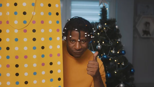 Kerstsfeer. Afro-Amerikaanse zwarte man met grote gouden boodschappentas een geschenk voor de kerstboom met duim omhoog — Stockfoto