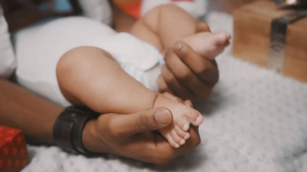 Niedliche Babyfüße in den Händen eines schwarzen Vaters, umgeben von Weihnachtsgeschenken, aus nächster Nähe — Stockfoto