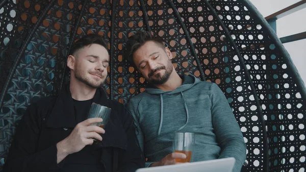 Νεαρό όμορφο γκέι ζευγάρι γελάει και χαλαρώνει στην κούνια με μπύρα. Βλέποντας ταινία στο λάπτοπ — Φωτογραφία Αρχείου