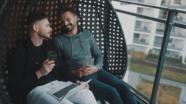 Молода красива гей-пара розслабляється на гойдалках на балконі з пивом і переглядом соціальних мереж на ноутбуці — стокове фото