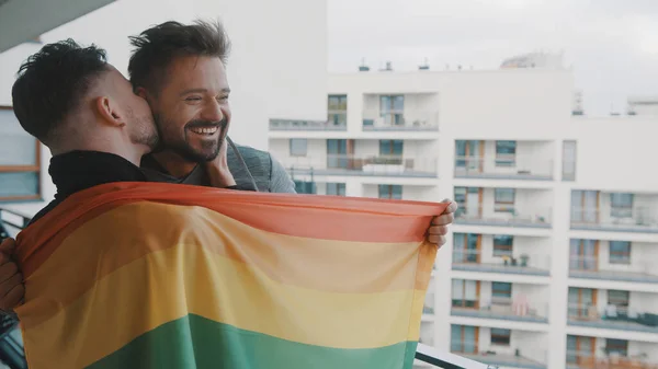 Pasangan muda gay yang bersemangat berdiri di balkon ditutupi dengan bendera pelangi. Seorang pria mencium suaminya — Stok Foto