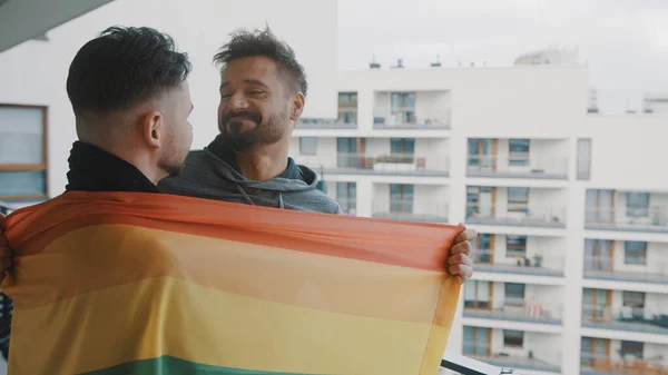 Jóvenes excitados lgbt pareja masculina de pie en el balcón cubierto con la bandera del arco iris — Foto de Stock