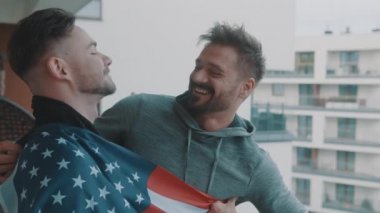 Heyecanlı yeni evli ve yakışıklı eşcinsel çift balkonda Amerikan bayrağı altında sarılıyorlar.