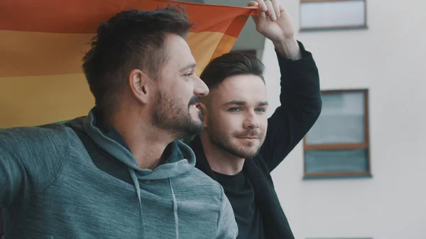 Jovem casal homossexual desfrutando da vista da varanda coberta com bandeira do arco-íris orgulho — Fotografia de Stock