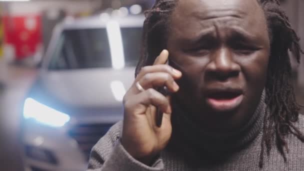 Hombre negro afroamericano molesto hablando en el teléfono inteligente delante del coche en el garaje — Vídeos de Stock