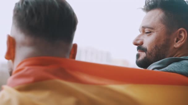 Bagsidebillede af homoseksuelle mandlige par dækket med regnbueflag krammer og smiler til hinanden – Stock-video