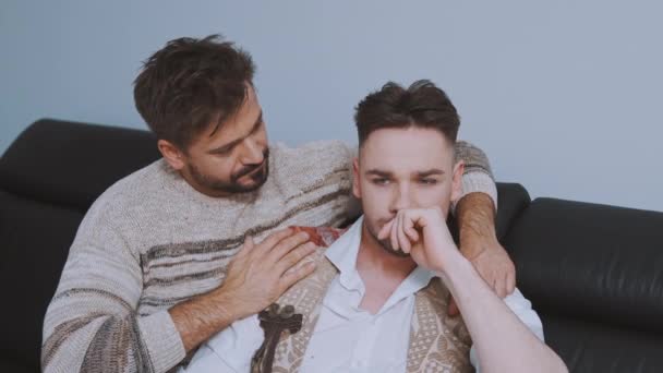 Молодые красивые белые мужчины, пара геев, у которых проблемы в отношениях сидят на диване и обнимаются — стоковое видео