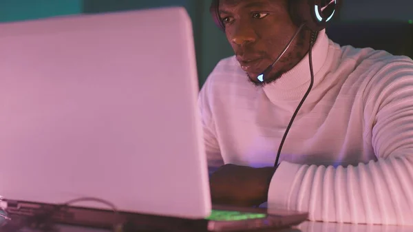 Hombre negro, adicto al trabajo con auriculares iluminados usando portátil por la noche —  Fotos de Stock