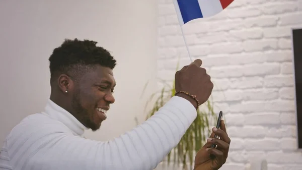 Zwarte man met een video-oproep of het kijken naar een spel op de smartphone tijdens het zwaaien vlag van Frankrijk — Stockfoto