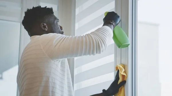 Um negro africano a limpar o apartamento. Pulverizar e limpar as molduras das janelas — Fotografia de Stock