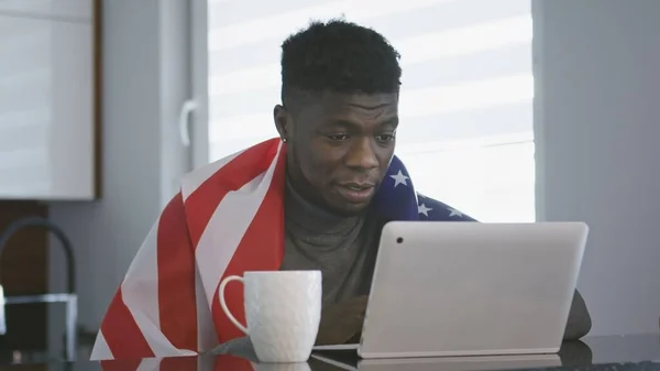アメリカを持つアフリカ系アメリカ人の男性は、テーブルの上にお茶を飲みながらノートパソコンで試合を見て肩にフラグを立てます。驚いた顔の表情 — ストック写真