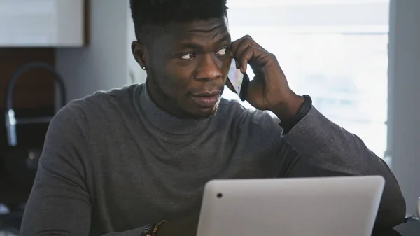 Trabajando remotamente. Africano americano negro hombre de negocios teniendo llamada telefónica en su apartamento mientras mira el portátil — Foto de Stock
