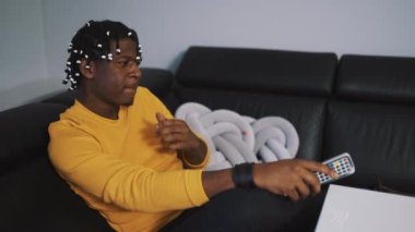 Genç Afro-Amerikan siyah adam elinde kumandayla evde televizyon seyrediyor.