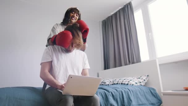 Молодая женщина отвлекает своего парня от работы на ноутбуке. Искатель внимания в боксёрских перчатках играет с партнерами длинными волосами — стоковое видео