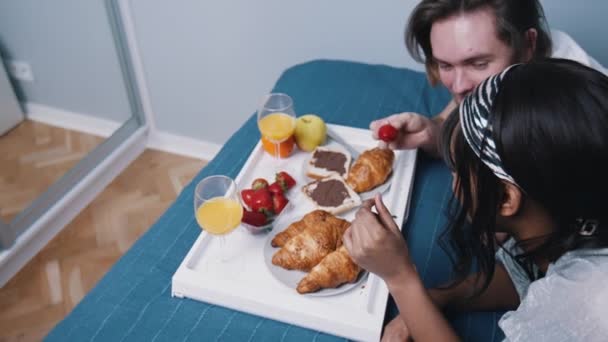 Jong multiraciaal koppel ontspannen in de slaapkamer en ontbijten op bed. Man die aardbeien aan zijn vriendin geeft — Stockvideo