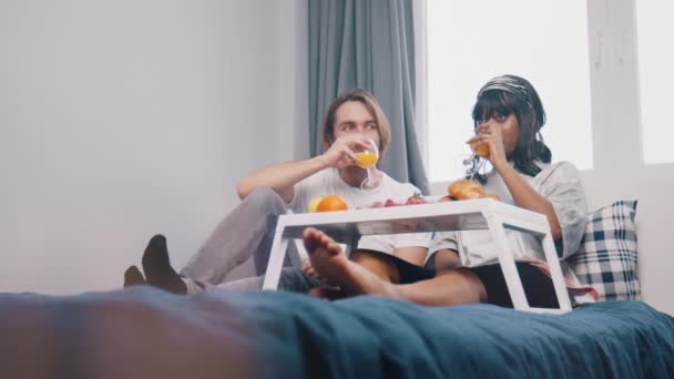 Ленивое утро в постели. Молодая многонациональная пара пьет сок и завтракает в спальне — стоковое видео