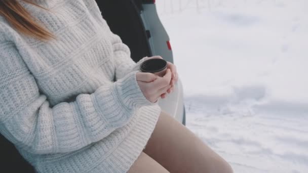 Крупним планом, молода жінка спирається на машину і п'є гарячий напій в зимовий сніжний день — стокове відео