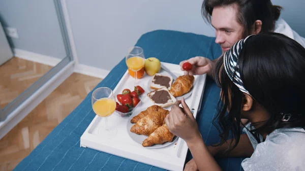 Jong multiraciaal koppel ontspannen in de slaapkamer en ontbijten op bed. Man die aardbeien aan zijn vriendin geeft — Stockfoto