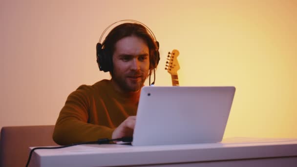 Egy fiatalember fejhallgatóval, aki laptopon dolgozik, vagy videokonferenciát tart. Gitár a háttérben — Stock videók