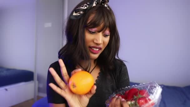 Beautiful young woman vlogging about diet. Holding orange and strawberries pack — Stockvideo