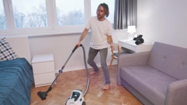 Evde elektrikli süpürgeyle dans eden beyaz bir genç.