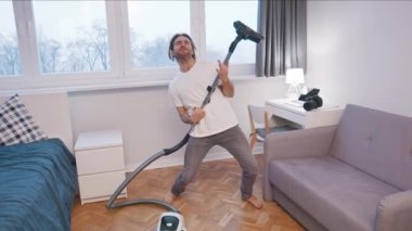 Genç beyaz adam ev işi yaparken eğleniyor. Dans eden şarkı söyleyen ve elektrik süpürgesini gitar olarak kullanan