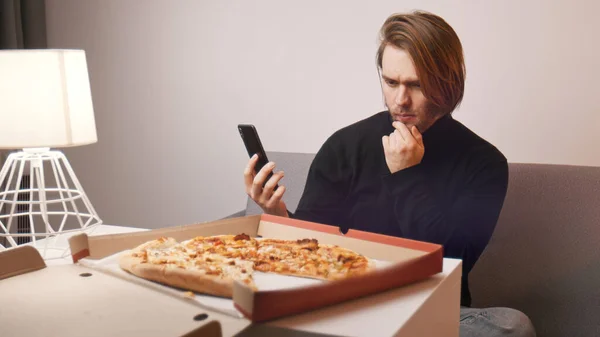 젊은 코카 시안 남성이 피자에 대해 불평하고 있습니다. 스마트폰을 들고 고객 지원을 요청하는 모습 — 스톡 사진