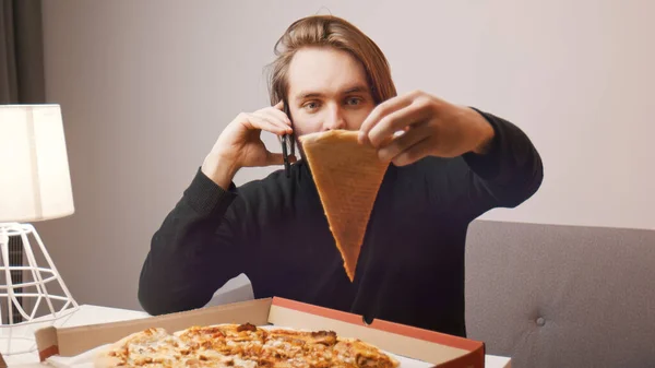 Młody biały mężczyzna narzeka na pizzę przez telefon. Trzymanie jednego kawałka podczas korzystania ze smartfona — Zdjęcie stockowe